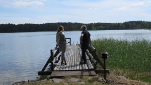 Henry Ahola från Träskända står tillsammans med fastighetsmäklare Stella Limnell på bryggan och njuter av utsikten. Kanske Ahola funderar om gammelgäddan lurar där i vassen. Eller så funderar han om stugan i Salo ändå var bättre. Två personer, en kvinna och en man, står på en lång brygga och tittar ut mot havet.