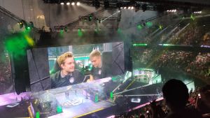 Jesse Vainikka ja OG-joukkueen kapteeni Johan "N0tail" Sundstein The Internationalin jättiruuduilla. En arena fylld med folk. I mitten syns en stor skärm som visar två män som ser emotionella ut.