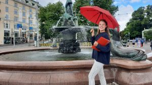 Maria Aho har jobbat som turistguide i mer än två år. Hennes guidningar börjar alltid från Havis Amanda i Helsingfors. Maria Aho står med ett rött paraply framför Havis Amanda.