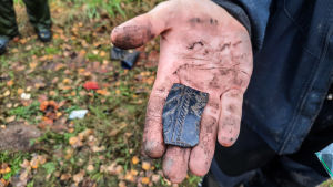 Arkeolog Taisto Palo visar upp en del av ett tennföremål som man hittat. En del av ett tennföremål i en hand. Den svarta skärvan har dekorationer på sig.