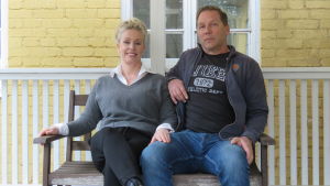 Annika Kalvia och Aki Karihtala pustar ut en stund utanför en av Bromarfhemmets flera ingångar. De har mestadels själva städat, målat och inrett det gamla äldreboendet, som är byggt 1925. Huset var i bra skick när det köptes, säger de båda. En kvinna och eman sitter på en bänk ute på en veranda. i bakgrunden ser man en gul yttervägg och ett större fönster.