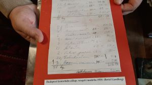 Originalreceptet till konfekten Budapest, handskrivet av Bertel Gardberg 1939. två händer håller i ett rött ark med vitt papper pålimmat. Den för hand skrivna texten är originalreceptet till konfekten Budapest