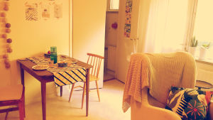 Ett brunt bord med rutig duk och två stolar bredvid. I förgrunden en vit fotölj med en mönstrad kudde.
