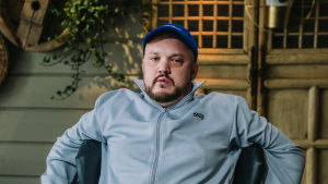 Rap-artisti Pyhimys istuu sisätiloissa Vain elämää -ohjelman kuvauksissa. Nojaa käsillään tuolin selkänojaan.