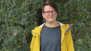 Susanna Kankare letar efter nya utmaningar nu när destilleriet är sålt. En kvinna med kort hår, glasögon och gul rock. Står framför en stor silverpil (ett träd).