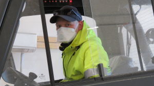 Jim Bäckman jobbar på Victor Ek i Hangö. Munskydd är obligatoriskt när han jobbar inne i den stora hallen, till och med då han sitter ensam i en truck. EN man inne i i hytten i en gaffeltruck. Han har neongula arbetskläder och keps, men det mest speciella är att han har munskydd. Det beror på coronaepidemin och regler på mannens arbetsplats.