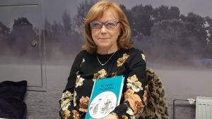 Professor emerita Maija Grönholm äldre dam i rött hår, svart blus och glasögon sitter med bok i famnen