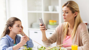 Mamma sitter och tittar på sin telefon vid matbordet, dotter tittar ledset på mamman