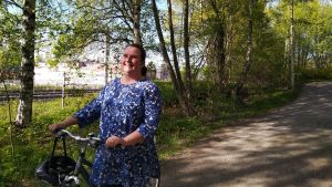 Päivi Metsäniemi anser att man borde bygga hus som passar in i Mosabackas miljö. En leende kvinna står med sin cykel på en sandväg. I bakgrunden finns gröna träd och järnvägsspåret skymtar.