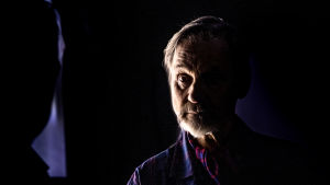 Asko Sarkola under repetitionerna av Pappan. Asko Sarkola fotograferad i ett mörkt rum i pjäsen "Pappan". Sarkola ser sig själv i spegeln och endast halva av hans ansikte syns medan den andra är täckt av mörkret.