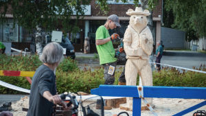 Kring tävlingsplatserna råder ett ständigt vimmel. Nainen pyörällä katsoo miestä joka veistää kulmahiomakoneella karhun mallista puuveistosta.