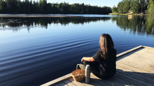 Coronaviruset har lett till att många har flyttat ut till sommarstugan - där ska man också få jobba tycker SFP. Ung kvinna sitter på en brygga vid Gallträsk i Grankulla med en korg och en termos bredvid sig.