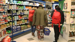 Korgen fylls med mat för resten av veckan. Mannen berättar ivrigt om vilka olika maträtter han ska bjuda sina vänner på. Tre personer inne i en matbutik. Bilden tagen bakifrån vid mjölkhyllan.