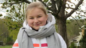 Längre sovmornar och mindre jäkt med att fixa sig klar till skolan eller jobbet. Det är nog inte bara Milla Hyvönen som har noterat den fördelen med den annars rätt tuffa coronavåren. En 15-årig flicka med blont hår i hästsvans bak. Hon har en vit, vindtät jacka och grå halsduk på sig. Hon ser in i kameran och ler lite. Ett träd och ett hus i bakgrunden.