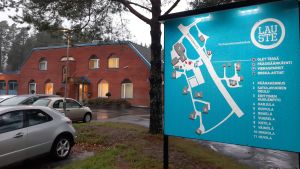 Familjerehabiliteringscentret har många olika verksamheter. En turkos skylt med karta över Lauste familjerehabiliteringscenter, och huvudbyggnaden i rödtegel i bakgrunden.