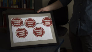 András Kádár visar upp de klistermärken som ungdomspartiet Fidelitasz lämnade efter sig när de demonstrerade utanför organisationens byggnad. Texten lyder: "Denna organisation stödjer invandring". András Kádár visar upp en inramad bild med de klistermärken regeringens ungdomsparti Fidelitasz lämnade efter sig.