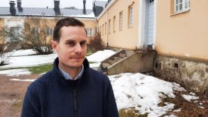 Gustav Ramberg vid den gamla sjukhusbyggnaden på Själö, som nu inrymmer forskningscenter, hotell och restaurang. Gustav Ramberg, en man med mörkt hår och blå tröja, står vid en stor, gammal, gul stenbyggnad en vinterdag.