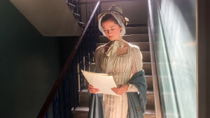 Jane Austen: Suljettujen ovien takana. Nainen seisoo portaikon alapäässä ja lukee papereita.