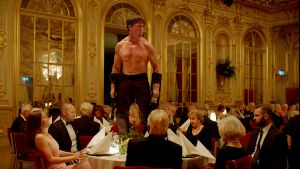 Storvuxen man utan skjorta står på ett middagsbord, de finklädda gästerna är förfärade.