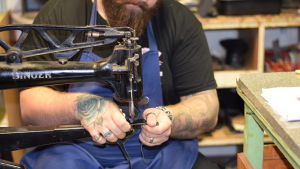 Det behövs ingen elektricitet, men nog en hel del övning för att sömmen ska bli rak och snygg. En man med tatuerade armar som syr i läder på en gammal symaskin. Man ser inte ansiktet på mannen.