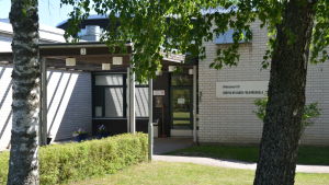 Efter midsommar riktar många blickarna hit. Ingång till vit tegelbyggnad. Träd och gräsmatta utanför.