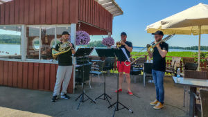 Bleckblåstrion började dagen med att spela nere vid Hammars strand. Därefter fortsatte de bland annat till Gammelbacka och Vårberga. En bleckblåstrio står och spelar på en uteservering.