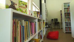 En hylla med barnböcker, andra hyllor med böcker i ett bibliotek, en röd säckdyna på golvet, i bakgrunden en dator på ett bord.
