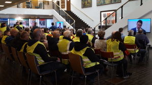 Publiken som följde med mötet via skärm i ämbetshusets aula var aktiv och återkom ofta med applåder. Människor i gula reflexvästar följer med fusionsdebatt i Korsholm.