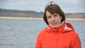 Anna-Karin Almén har forskat i hoppkräftor sedan 2012 och hoppas att hon kan fortsätta att forska i hur klimatförändringar påverkar havet. En kvinna i orange jacka och blå byxor står på en strand med vågor som rullar in. Hon heter Anna-Karin Almén.