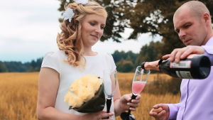 Ung man och kvinna i bröllopskläder står på äng och häller upp champagne i glas