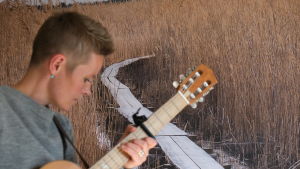 Musik från skärgården. Désirée Saarela med gitarr i profil, i bakgrunden en bild av vass och en träspång.