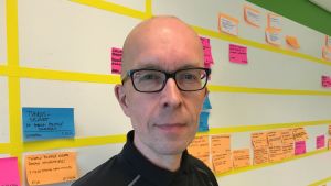 Mies kasvokuvassa, taustalla kanban-seinä ja tehtävälappuja.
