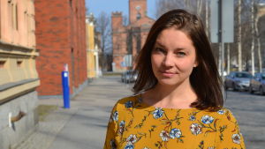 Cecilia Flygar. En ung kvinna med långt mörkt hår står ute på en gata. I bakgrunden syns en röd tegelkyrka.