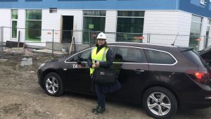 Terhi Halla utanför ett av höghusen i Nickby medan byggandet ännu var på hälft. En svartklädd kvinna i neongul skyddsväst och vit hjälm står framför en svart bil som är parkerad framför ett höghus som håller på att byggas.