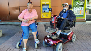 Tua Sjöholm och Helmer Wiik har svårt att tro att Sibboborna mår bättre än någon annan. En äldre kvinna och en äldre man utanför en matbutik. Mannen sitter på en elskoter och kvinnan på en rullator.