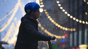 Det är främst för gående, cyklister och andra i trafiken som inte tar sig fram med bil, som ljuden i de låga hastigheterna behövs. En elsparkcykelåkare med blå hjälm står i skymningen i regn vid Alexandersgatans julbelysning i Helsingfors.