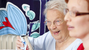 En äldre kvinna som putsar ett silverföremål, en annan kvinna (vårdpersonal) syns i förgrunden.