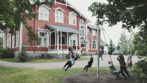 Suuri punainen koulurakennus valkoisine ikkunanpuitteineen. Etualalla pihalla lapset pyörivät narujen varassa tolppaa ympäri.
