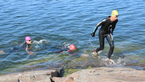 Swimrun kombinerar simning och löpning med naturupplevelse. En man iklädd våtdräkt tar sig upp ur havet längs med klippor. Två andra män väntar på sin tur.