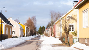 Husen längs den här gatan är alla från samma årtionde, men kreosot finns ingalunda i alla hus. Färggranna trähus längs snöig gata i solsken.