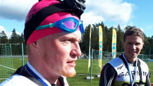I mål, trötta, men nöjda, trots lite bister min. Sebastian Dahlström dokumenterade sitt första swimrun med en kamera på pannan. Till höger parhästen Niclas Wiitala. Två män i våtdräkt med medaljer runt halsen, den ena har dessutom en neonrosa simmössa. De är på ett sportplan. Blå himmel och sol. September 2020.