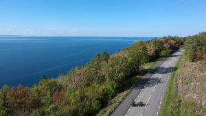 Utan försäkring men glada ändå. En bild tagen uppifrån luften, med drönare. Till vänster syns blått hav. Till höger två cyklister som kommer cyklande på en landsväg. Träden är gröna och rostbruna. Det börjar bli höst.