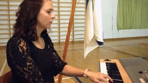 Rektor Jennie Ahtola anställdes vid årsskiftet och kom direkt in i jubileumsåret som uppmärksammats på flera olika sätt. Hon ackompanjerade också sångerna som sjöngs under festdagen. En kvinna sitter vid en elorgel och spelar i en skolas gymnastiksal. Bakom henne finns också Finlands flagga.