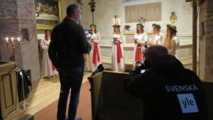 Kjell Wikström från Ekenäsnejdens församling filmar luciakröningen i Tenala kyrka. Yle Västnylands Christoffer Westerlund gör i sin tur en video på hur årets luciakröningar fixas i år. En man bandar lucia och tärnor inne i en kyrka, Yles tekniker filmar i sin tur mannen som filmar. Båda männen har munskydd.