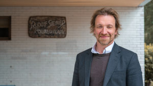 Marco Mäkinen Helsingin Rudolf Steiner -koululla Kuvassa on Marco Mäkinen Helsingin Rudolg Steiner -koulun ulkoseinän edessä.Vieressä näkyy koulun kyltti.