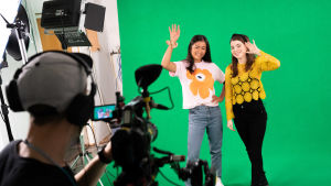 Kaksi naista heiluttamassa kameralle studiossa vihreän taustan edessä.