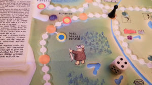 När spelaren väl kommer i mål har hen förtjänat sin vargsvans. Sluga Sibbe ger den dock inte utan att få en bra slant i utbyte. Närbild på brädspelet Sibbo-vargen med en tärning och en spelpjäs. På bilden syns målet där en viking väntar med en vargsvans i handen.