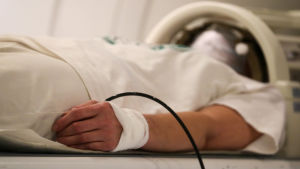Potilas makaa laitteessa, jossa kuvataan päätä. Kädessä on sideharsoa, josta lähtee jokin letku.