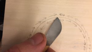Dubbla rader med pilar i en cirkel, skissade i blyerts på en ljus träskiva och en hand som håller en kniv med kort egg, som används för karvsnitt.