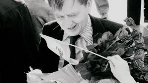 Åsikterna om Olof Palme var delade bland svenskarna under hans många år som statsminister. Svartvit bild av Olof Palme med en bukett rosor och kampanjvimpel.